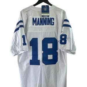 Może przedstawiać: Biała koszulka piłkarska z niebieskimi i białymi paskami na rękawach. Z tyłu koszulki widnieje nazwisko "MANNING" i numer "18" w kolorze niebieskim.