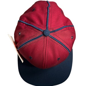 May include: A red baseball cap with a navy blue brim and a navy blue stitching detail. The cap has a button on top.