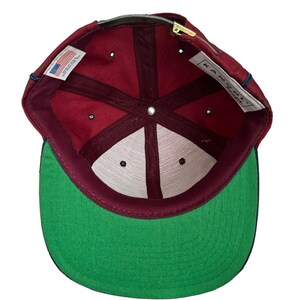 May include: A maroon baseball cap with a green underside and a navy blue brim. The inside of the cap has a white label with the text "KAN-C-ON" and a small American flag label.