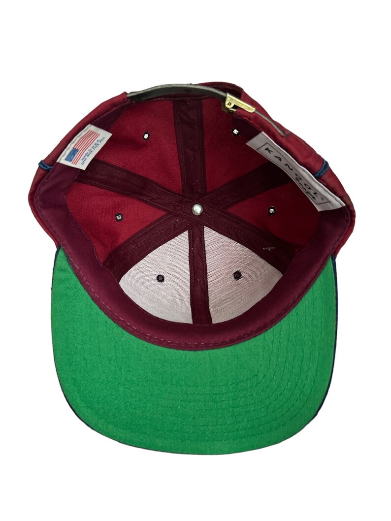 May include: A maroon baseball cap with a green underside and a navy blue brim. The inside of the cap has a white label with the text "KAN-C-ON" and a small American flag label.