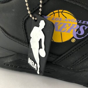 Puede incluir: Zapatillas de baloncesto de cuero negro con un logotipo de Los Angeles Lakers amarillo y morado y una cadena plateada con un dije de logotipo de la NBA en blanco y negro.