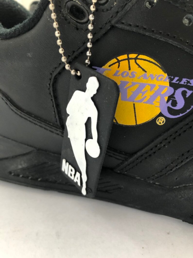 Puede incluir: Zapatillas de baloncesto de cuero negro con un logotipo de Los Angeles Lakers amarillo y morado y una cadena plateada con un dije de logotipo de la NBA en blanco y negro.
