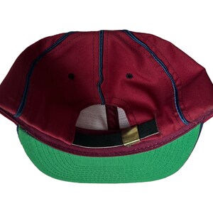May include: A red baseball cap with a green brim and a black strap. The cap has a navy blue stitching detail.