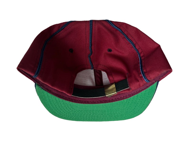May include: A red baseball cap with a green brim and a black strap. The cap has a navy blue stitching detail.