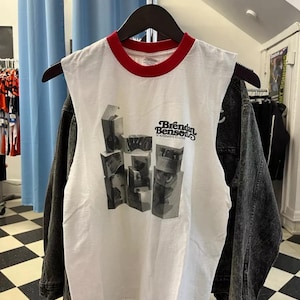 May include: White sleeveless t-shirt with a red trim around the collar. The shirt features a black and white graphic design and the text "Brendan Benson" with "An Alternative to Love" below it. A black denim jacket hangs behind the shirt.