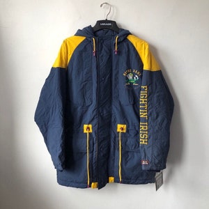 May include: A blue and yellow hooded jacket with a Notre Dame Fighting Irish logo on the front. The jacket has a zipper closure and two pockets on the front.