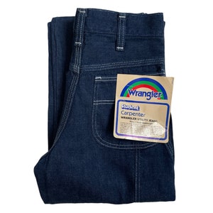 以下が含まれることがあります： 白いステッチが入った濃い青色のデニムジーンズ。ジーンズには「Wrangler Student Carpenter Wrangler Utility Jeans Full 1 Year Warranty」と書かれたラベルが付いています。