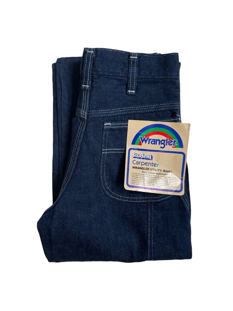 以下が含まれることがあります： 白いステッチが入った濃い青色のデニムジーンズ。ジーンズには「Wrangler Student Carpenter Wrangler Utility Jeans Full 1 Year Warranty」と書かれたラベルが付いています。