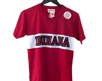 Könnte beinhalten: Rotes T-Shirt mit kurzen Ärmeln, einem weißen Streifen über der Brust und dem Wort "INDIANA" in schwarzen Buchstaben. Das Shirt hängt an einem schwarzen Kleiderbügel.