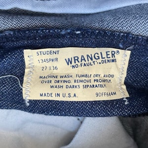 May include: A close-up of a blue denim Wrangler "No-Fault" denim label with white stitching. The label has the text "Student 134SPWR 27 x 36" and care instructions. The label is made in the U.S.A.