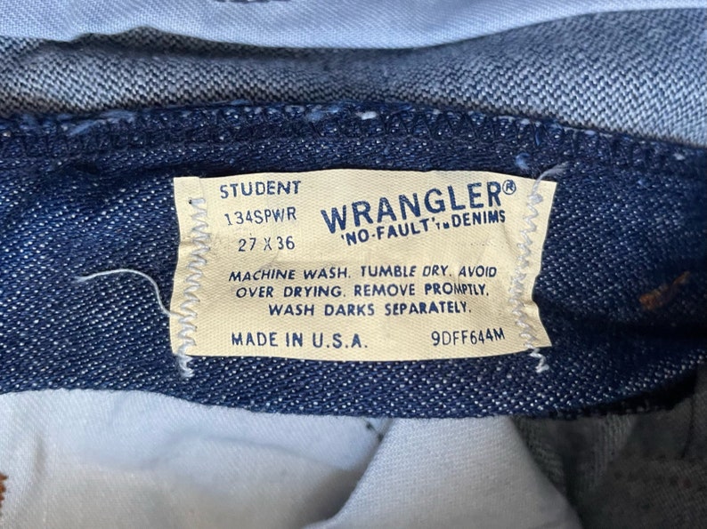 May include: A close-up of a blue denim Wrangler "No-Fault" denim label with white stitching. The label has the text "Student 134SPWR 27 x 36" and care instructions. The label is made in the U.S.A.