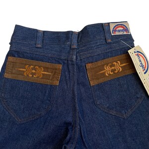May include: A pair of blue denim jeans with brown corduroy patches on the back pockets. The patches have an embroidered design. The jeans have a Wrangler label on the back waistband and a tag that says "No-Fault 100% Cotton Denim with "Tailor" Fit."