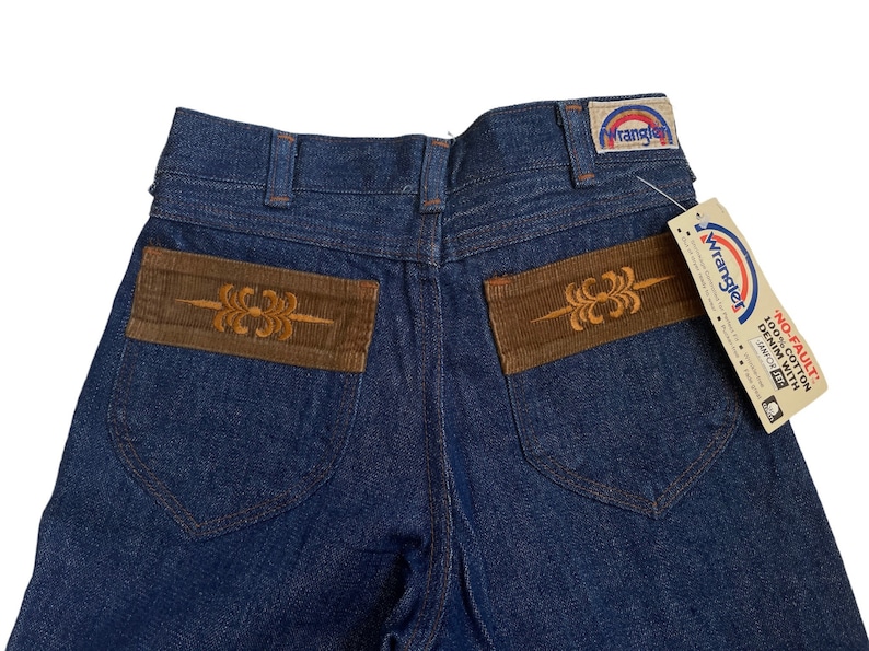 May include: A pair of blue denim jeans with brown corduroy patches on the back pockets. The patches have an embroidered design. The jeans have a Wrangler label on the back waistband and a tag that says "No-Fault 100% Cotton Denim with "Tailor" Fit."