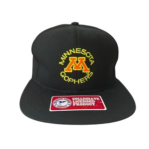 Puede incluir: Gorra de béisbol negra de ala plana con bordado dorado y amarillo "MINNESOTA GOPHERS". La gorra tiene una etiqueta de producto con licencia universitaria.
