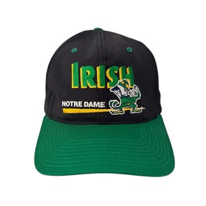 Op de afbeelding: Zwarte en groene baseballpet met de woorden "Irish Notre Dame" en een cartoon leprechaun mascotte.