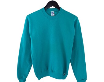 May include: A teal blue sweatshirt with a white logo on the front and a white tag on the sleeve. The sweatshirt is hanging on a black hanger.