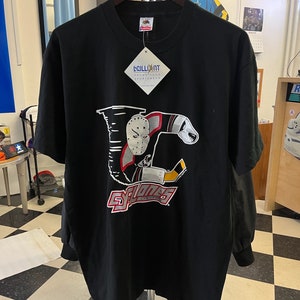 Può includere: Maglietta nera con una grafica rossa, bianca e nera di un giocatore di hockey con una maschera e un bastone da hockey. La grafica include il testo "Cyclones Cincinnati".