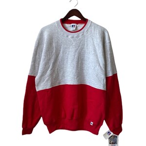May include: A gray and red sweatshirt with a red trim around the neck. The sweatshirt has a Russell Athletic logo on the left chest and a tag on the bottom right side. The tag has the Russell Athletic logo and the words "Russell Athletic" on it.
