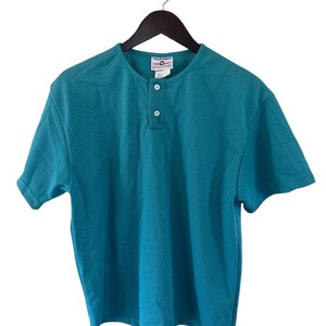 Op de afbeelding: Tealkleurig sportshirt met korte mouwen, een henley-halslijn en twee witte knopen. Het shirt is gemaakt van een ademende mesh-stof en hangt aan een zwarte hanger. Het label luidt "TEAMWORK ATHLETIC APPAREL".