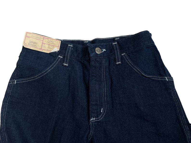 以下が含まれることがあります： 白いステッチと「3004PH-F CARPENTER PANTS 21"x30" MADE IN HONG KONG」と書かれた小さな紙タグが付いた濃い青色のデニムのカーペンターパンツ。