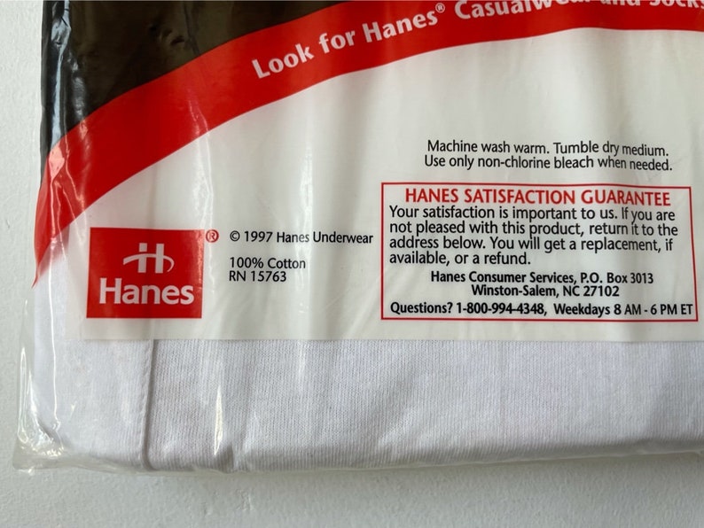以下が含まれることがあります： 白いHanesの下着のパッケージ。パッケージには、赤いHanesのロゴ、「100％コットン」の文字、「Hanes Satisfaction Guarantee」の文字が含まれています。 パッケージは透明なプラスチックで密封されています。
