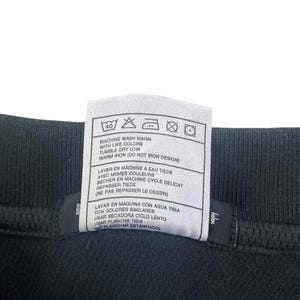 K&ouml;nnte beinhalten: Ein wei&szlig;es Pflegeetikett mit schwarzem Text und Symbolen. Das Etikett enth&auml;lt Anweisungen zum Waschen, Trocknen und B&uuml;geln eines Kleidungsst&uuml;cks. Der Text enth&auml;lt "Machine wash warm with like colors", "Tumble dry low", "Warm iron (Do not iron design)", "Laver en machine a eau tiede avec memes couleurs", "Secher en machine cycle delicat", "Repasser tiede (Ne pas repasser le dessin)", "Lavar en maquina con agua tibia con colores similares", "Usar secadora ciclo lento", "Usar plancha tibia (No planchar estampado)".