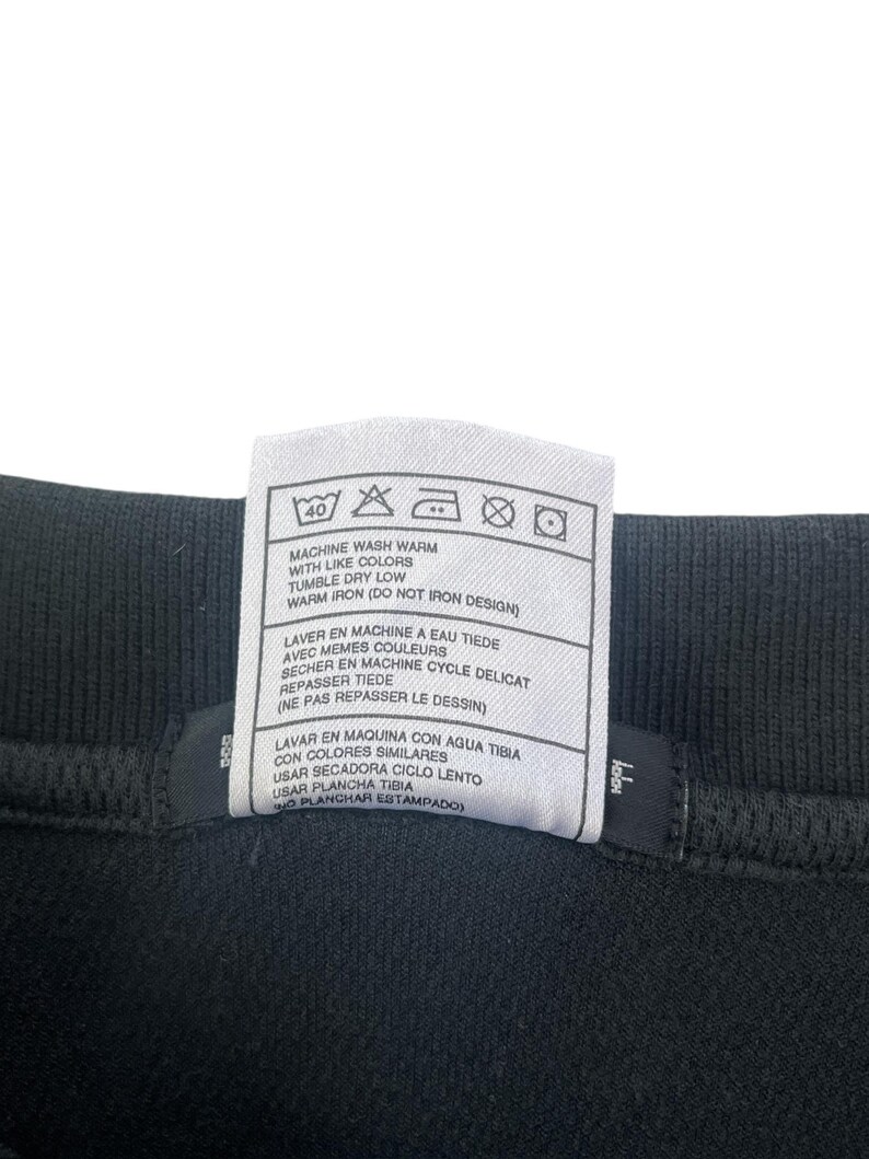 K&ouml;nnte beinhalten: Ein wei&szlig;es Pflegeetikett mit schwarzem Text und Symbolen. Das Etikett enth&auml;lt Anweisungen zum Waschen, Trocknen und B&uuml;geln eines Kleidungsst&uuml;cks. Der Text enth&auml;lt "Machine wash warm with like colors", "Tumble dry low", "Warm iron (Do not iron design)", "Laver en machine a eau tiede avec memes couleurs", "Secher en machine cycle delicat", "Repasser tiede (Ne pas repasser le dessin)", "Lavar en maquina con agua tibia con colores similares", "Usar secadora ciclo lento", "Usar plancha tibia (No planchar estampado)".