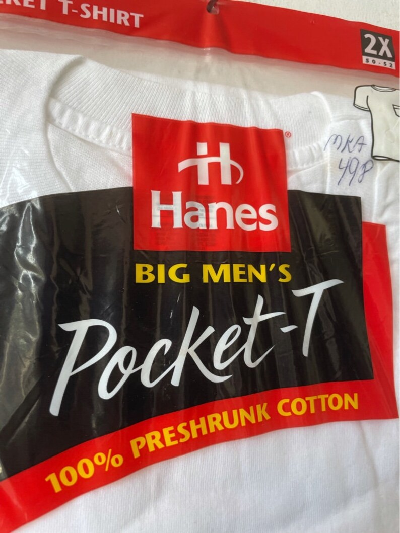 以下が含まれることがあります： 白いHanes Pocket-Tシャツがビニール袋に入っています。パッケージは赤、黒、白で、Hanesのロゴと「BIG MEN'S Pocket-T」の文字が印刷されています。シャツのサイズは2X、50-52です。