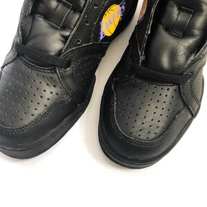 Puede incluir: Un par de zapatillas de baloncesto de cuero negro de ca&ntilde;a alta con un logotipo "Lakers" morado y amarillo en una zapatilla.