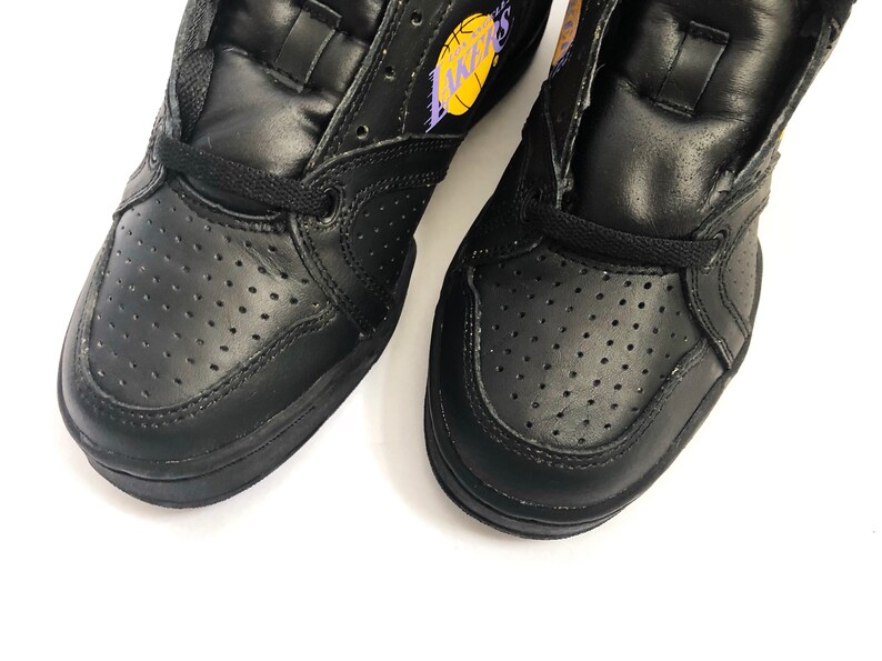 Puede incluir: Un par de zapatillas de baloncesto de cuero negro de ca&ntilde;a alta con un logotipo "Lakers" morado y amarillo en una zapatilla.