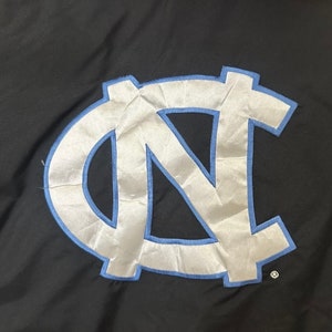 May include: Black jacket with a large, light blue and white embroidered logo. The logo features the letters "NC" in a stylized design, outlined in light blue. The jacket is made of a smooth, possibly nylon, material.