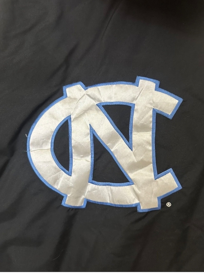 May include: Black jacket with a large, light blue and white embroidered logo. The logo features the letters "NC" in a stylized design, outlined in light blue. The jacket is made of a smooth, possibly nylon, material.