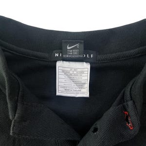 K&ouml;nnte beinhalten: Schwarzes Golf-Shirt mit einem Nike Golf-Logo auf der Vorderseite. Das Etikett lautet "RH# 56323 CA# 05553 REC NHE 920220 KL4" und "07-NAY CA 00663 IFC NME 900220 KLA 70% BAUMWOLLE 20% POLYESTER 10% ALGODON 10% POLYESTER HERGESTELLT IN THAILAND PFLEGEANLEITUNG AUF DER R&Uuml;CKSEITE".