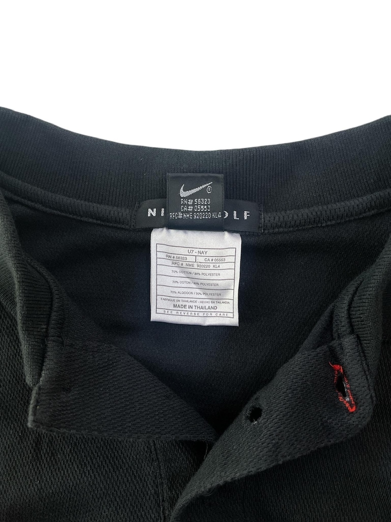 K&ouml;nnte beinhalten: Schwarzes Golf-Shirt mit einem Nike Golf-Logo auf der Vorderseite. Das Etikett lautet "RH# 56323 CA# 05553 REC NHE 920220 KL4" und "07-NAY CA 00663 IFC NME 900220 KLA 70% BAUMWOLLE 20% POLYESTER 10% ALGODON 10% POLYESTER HERGESTELLT IN THAILAND PFLEGEANLEITUNG AUF DER R&Uuml;CKSEITE".