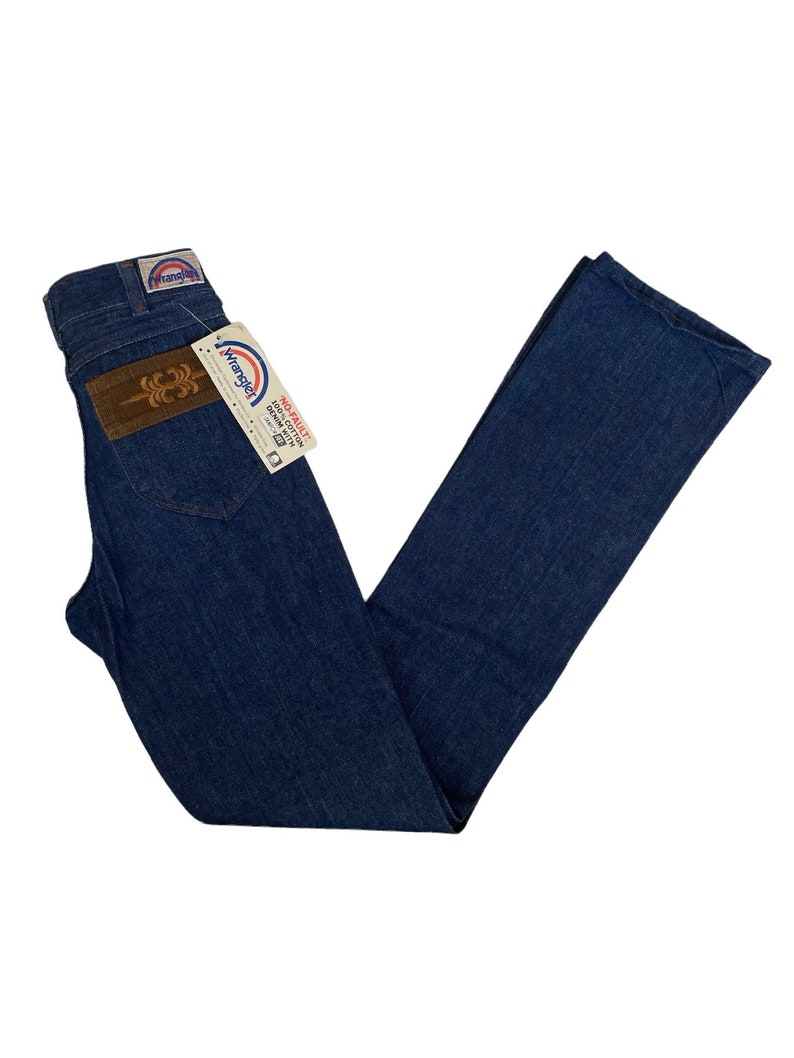 May include: A pair of blue denim jeans with a brown leather patch on the back pocket. The jeans have a rainbow-colored Wrangler label attached to the back pocket.