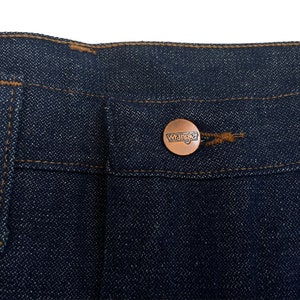 以下が含まれることがあります： 濃い青色のデニム生地のクローズアップ。生地には「Wrangler」と書かれた銅色のボタンが付いている。