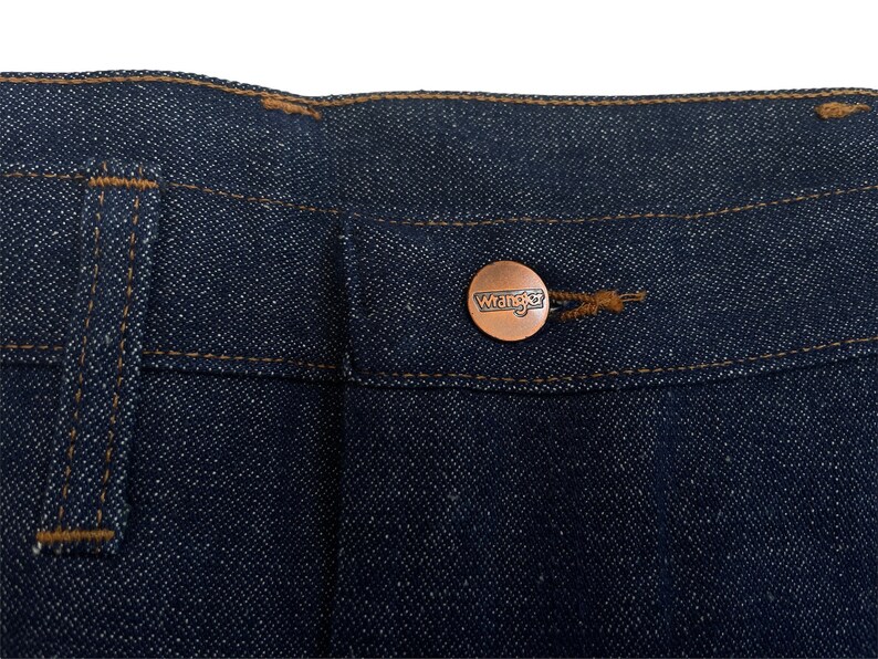 以下が含まれることがあります： 濃い青色のデニム生地のクローズアップ。生地には「Wrangler」と書かれた銅色のボタンが付いている。