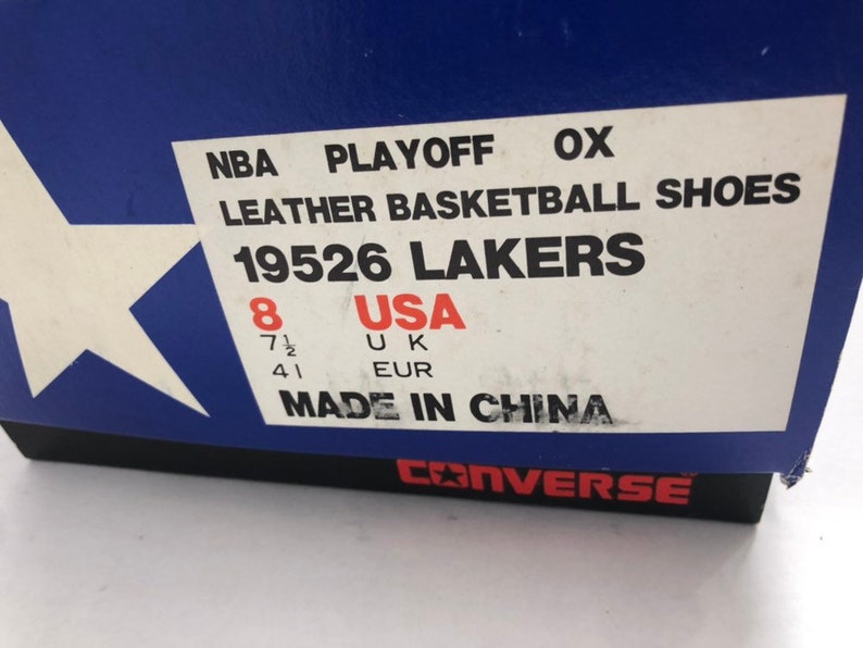 Puede incluir: Una caja blanca con ribete azul y una etiqueta blanca. La etiqueta dice "NBA PLAYOFF OX LEATHER BASKETBALL SHOES 19526 LAKERS" y enumera el tama&ntilde;o del zapato como "8 USA 7 1/2 UK 41 EUR". La etiqueta tambi&eacute;n dice "MADE IN CHINA". La parte inferior de la caja dice "CONVERSE" en letras negras.