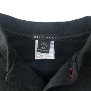 K&ouml;nnte beinhalten: Schwarzes Nike Golf Poloshirt mit einem wei&szlig;en Etikett mit der Aufschrift "Nike Golf" und "Hergestellt in Thailand". Das Etikett hat auch ein Gr&ouml;&szlig;enetikett mit einem "L" und einem "GG" in einem Kreis.