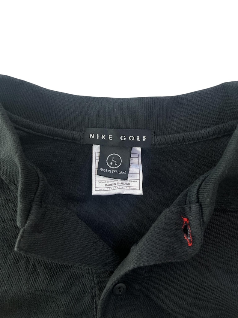 K&ouml;nnte beinhalten: Schwarzes Nike Golf Poloshirt mit einem wei&szlig;en Etikett mit der Aufschrift "Nike Golf" und "Hergestellt in Thailand". Das Etikett hat auch ein Gr&ouml;&szlig;enetikett mit einem "L" und einem "GG" in einem Kreis.