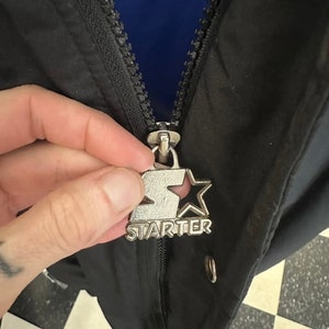 May include: Close-up of a black jacket with a silver zipper pull featuring the "Starter" logo. The jacket has a blue interior lining. The zipper is partially closed, revealing the jacket's construction and design.