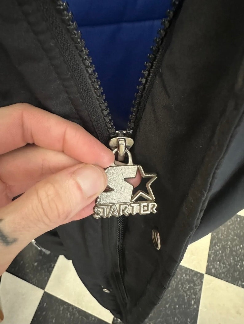 May include: Close-up of a black jacket with a silver zipper pull featuring the "Starter" logo. The jacket has a blue interior lining. The zipper is partially closed, revealing the jacket's construction and design.