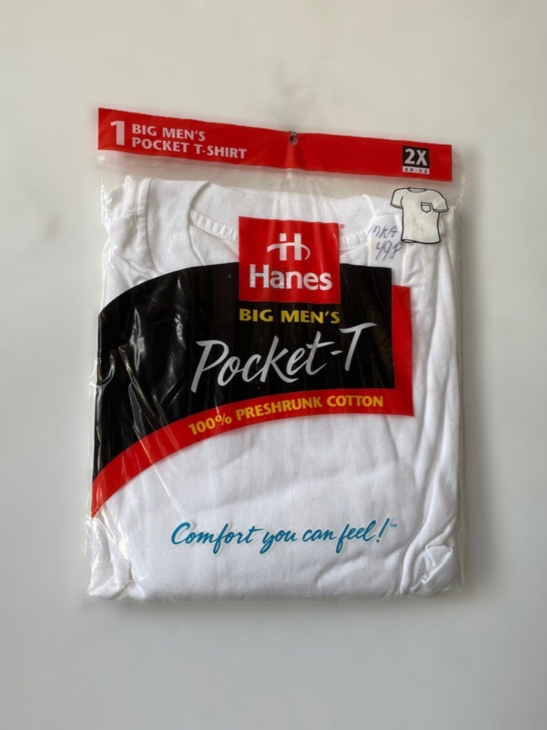 以下が含まれることがあります： パッケージされた白いHanes Pocket-Tシャツ。透明なプラスチックパッケージには、赤と黒のラベルが付いています。ラベルには「100％防縮加工コットン」と「Comfort you can feel！」と書かれています。シャツのサイズは2XLです。
