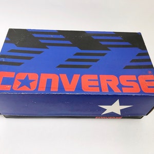 Puede incluir: Una caja de zapatos azul y negra con la palabra "CONVERSE" en letras rojas y un logotipo de estrella blanca. La caja tiene un fondo azul con rayas negras.