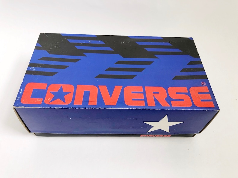 Puede incluir: Una caja de zapatos azul y negra con la palabra "CONVERSE" en letras rojas y un logotipo de estrella blanca. La caja tiene un fondo azul con rayas negras.
