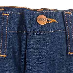 May include: Close-up of the waistband of a pair of blue denim jeans with a brown leather Wrangler label. The jeans have orange stitching.