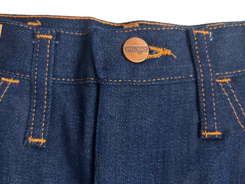 May include: Close-up of the waistband of a pair of blue denim jeans with a brown leather Wrangler label. The jeans have orange stitching.