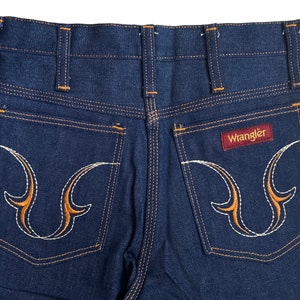 May include: A pair of blue denim jeans with orange stitching and a Wrangler logo on the back pocket. The back pockets have a unique embroidered design.