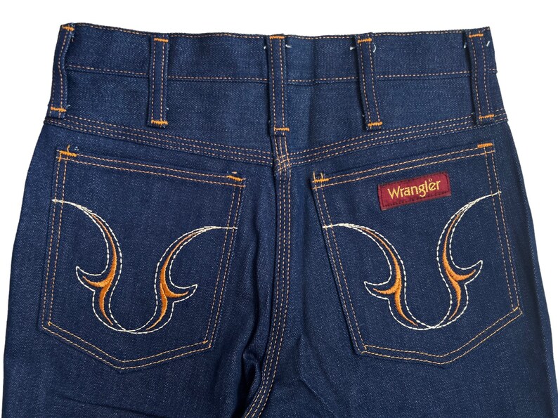 May include: A pair of blue denim jeans with orange stitching and a Wrangler logo on the back pocket. The back pockets have a unique embroidered design.