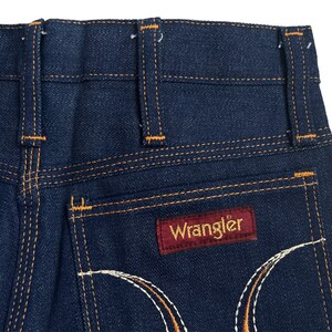 May include: A pair of dark blue denim jeans with orange stitching and a red leather Wrangler label on the back pocket.